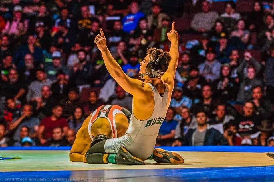 Two wrestlers on a mat, one raising both arms in celebration before a cheering crowd