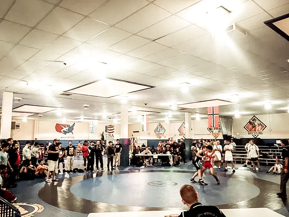 Wrestling match in a gymnasium, with two athletes on a mat and spectators around the ring.