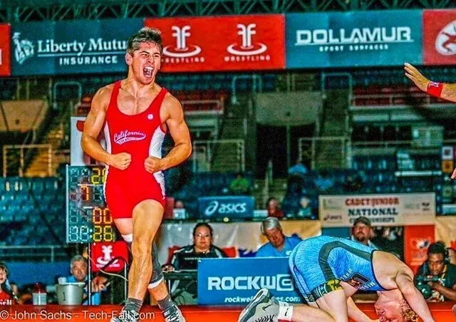 Two wrestlers compete on a mat, one in red standing over a blue-clad opponent in a stadium setting.