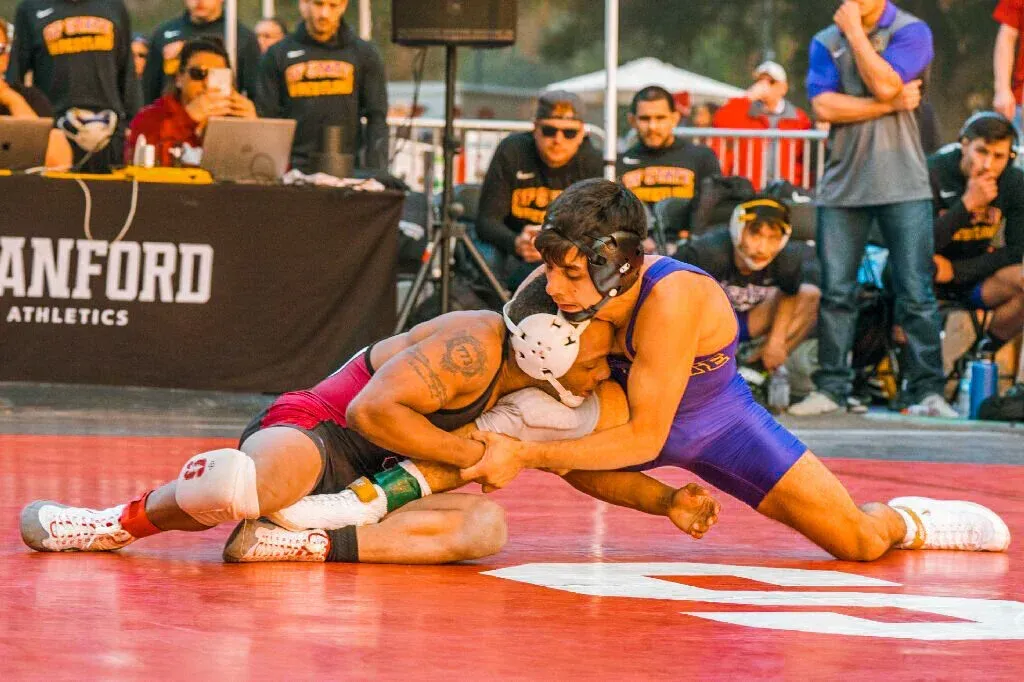 Two wrestlers in red and purple grapple on a mat during a Stanford athletics match.