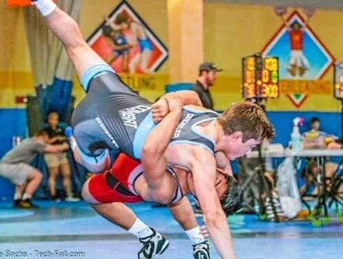 Two wrestlers grappling on a blue mat in a brightly lit indoor gymnasium