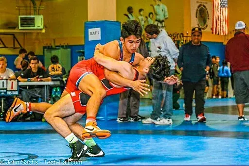 Two wrestlers in orange singlets grapple on a blue mat in a gymnasium.