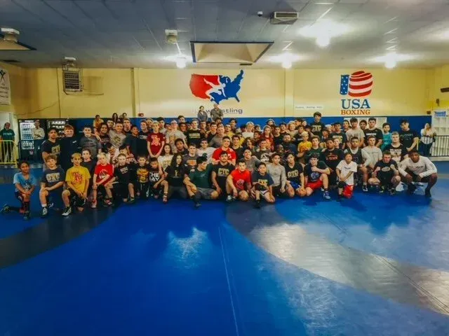 Large group photo in a martial arts gym with people posing on blue mats under USA banners