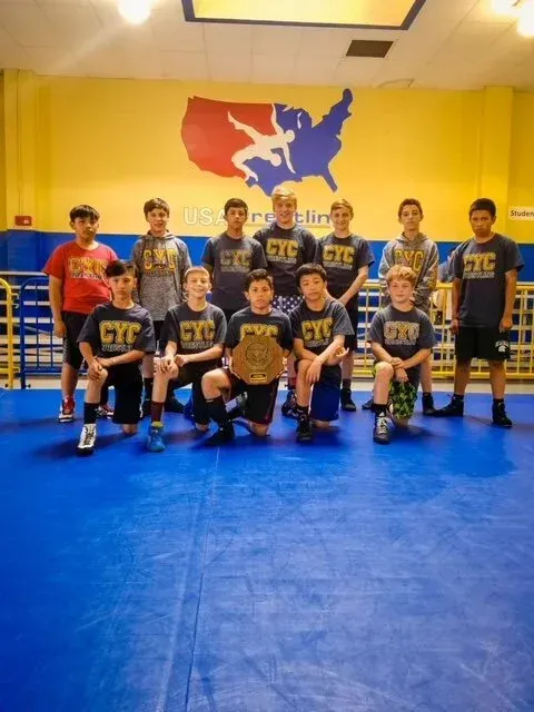 Youth sports team posing indoors on a blue mat, wearing matching black and yellow uniforms.