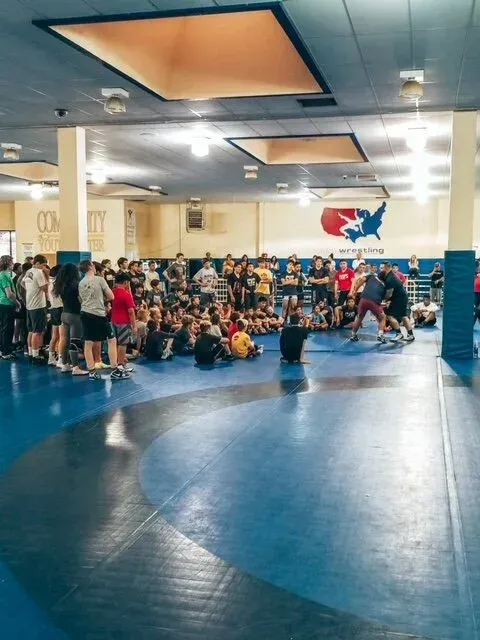 People seated in a wrestling gym with blue mats and a Texas map on the wall