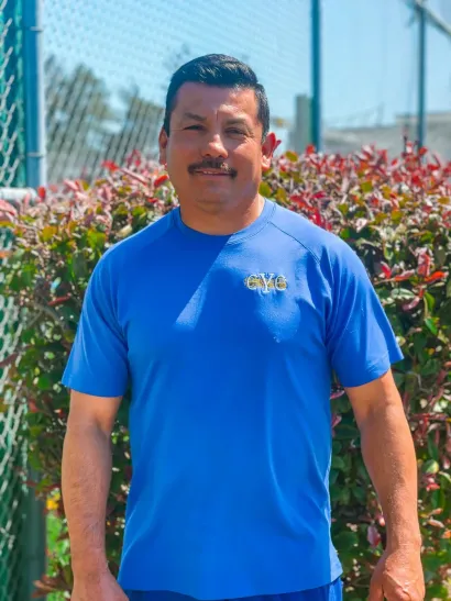 Man standing outdoors in a bright blue shirt in front of flowering bushes and a chain-link fence
