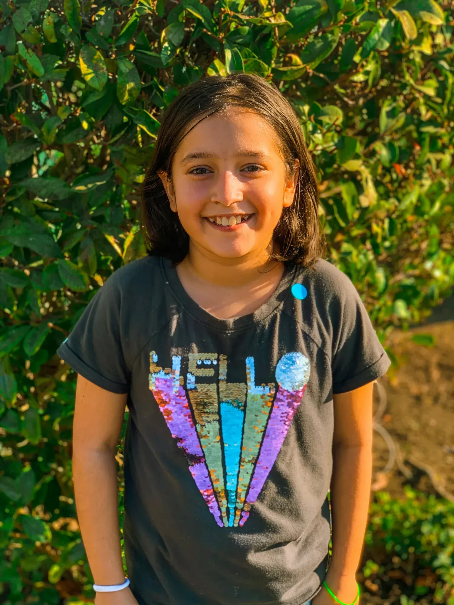 Smiling child standing outdoors in front of leafy green plants, wearing a dark T-shirt with colorful graphics.