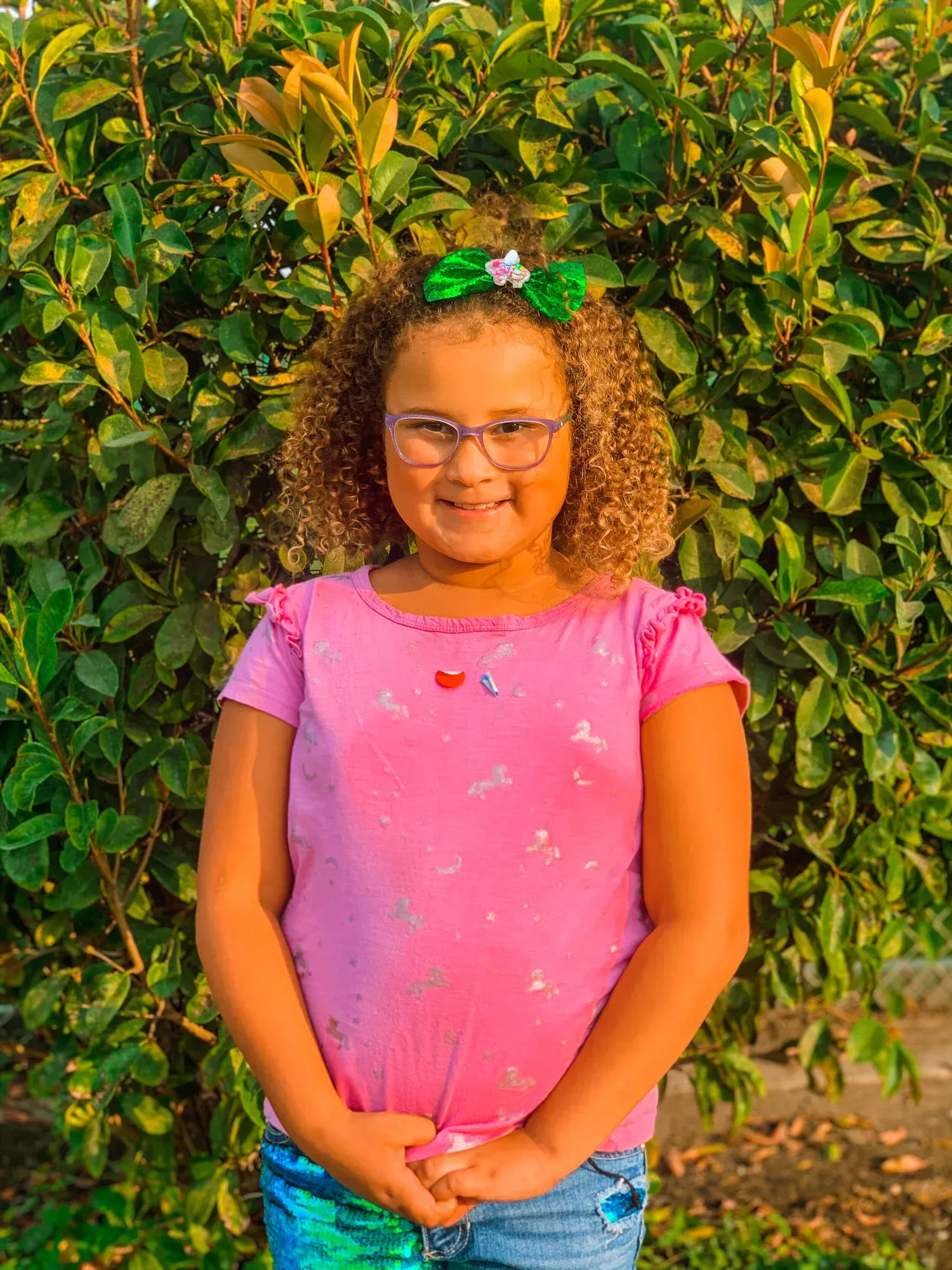 Smiling child in a pink shirt standing before a leafy green hedge