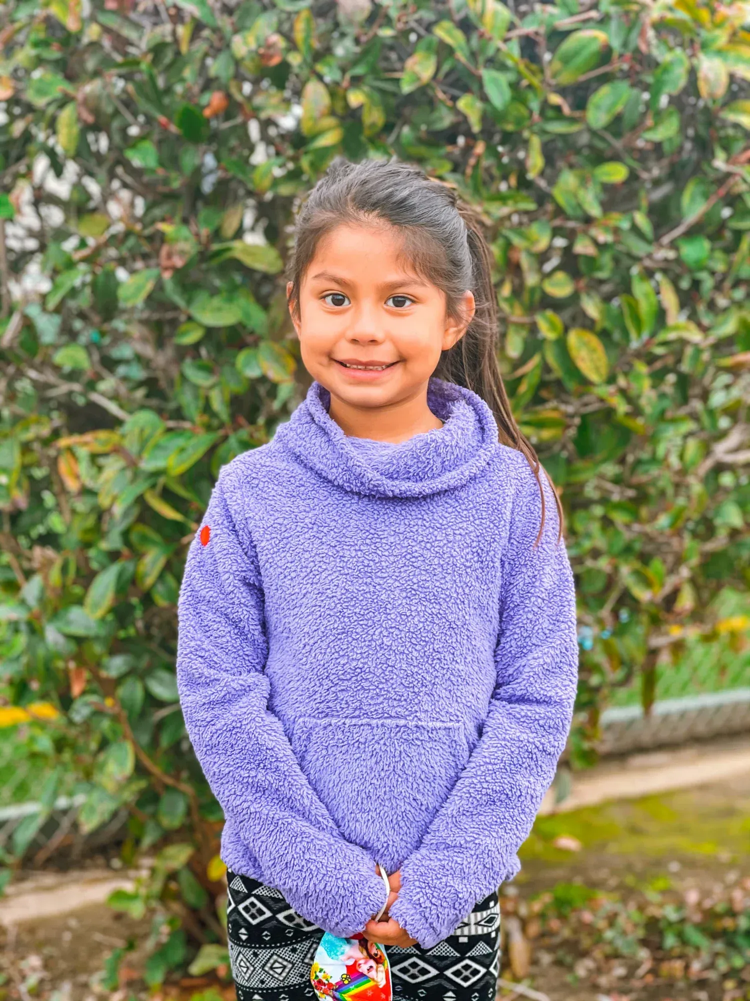 Smiling child in a purple sweater standing outdoors in front of green bushes