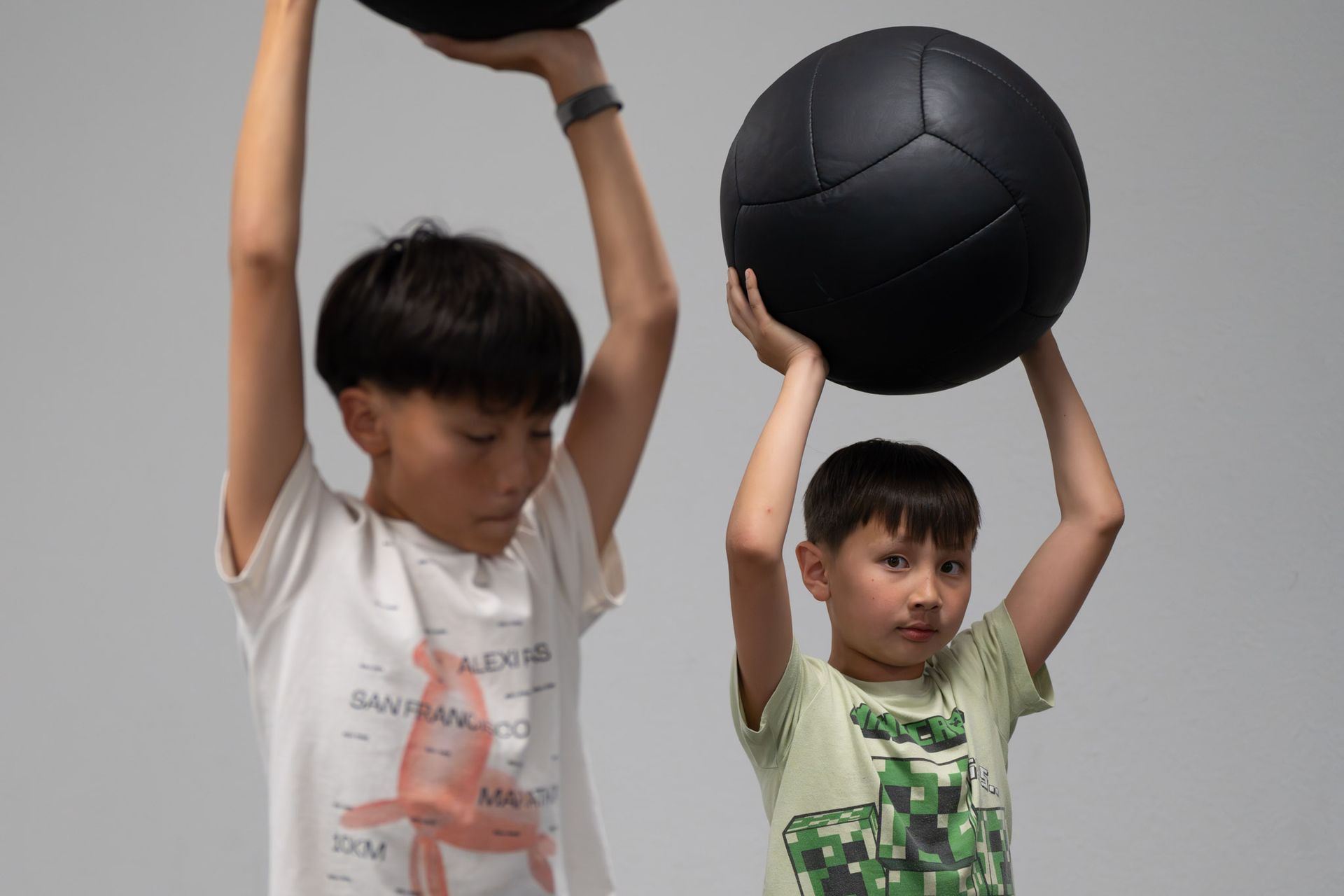 Two children lifting large exercise balls overhead against a plain gray wall
