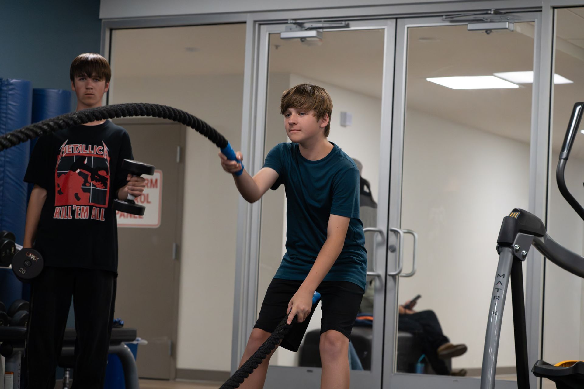 Two boys using battle ropes in a gym, with mirrors and exercise equipment behind them.