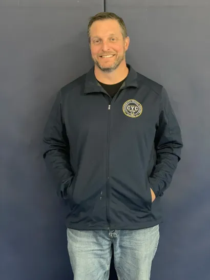 Man standing against a dark wall, wearing a navy jacket and jeans, with hands in pockets and smiling.