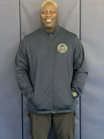 Man standing against a dark backdrop, smiling in a navy jacket and dark pants with hands in pockets.