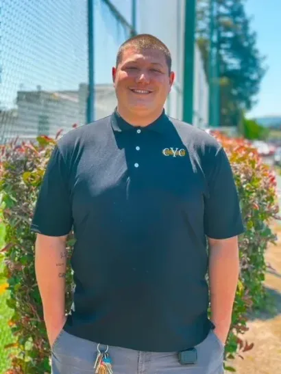 Man standing outdoors in a black polo shirt and jeans, smiling in front of flowers and a building.