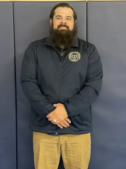 Man standing against a blue wall, wearing a dark jacket and khaki pants, hands clasped in front.