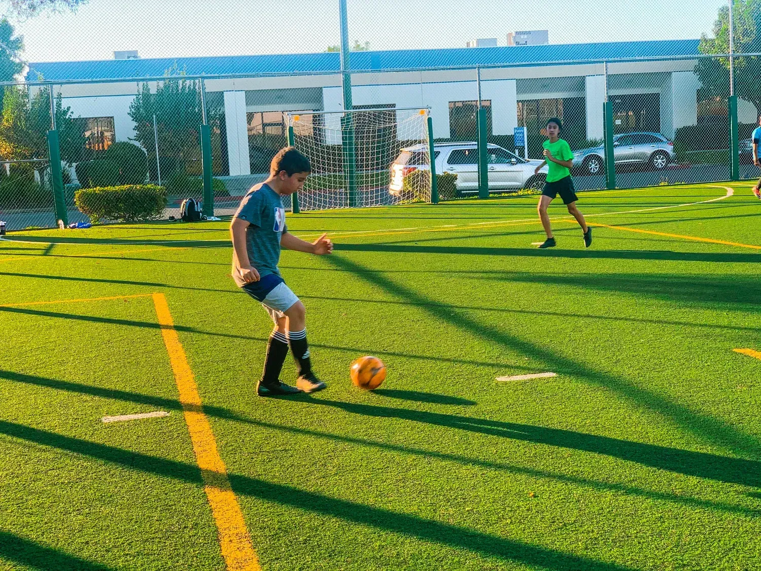 Children playing soccer on a sunny field, with one dribbling an orange ball near yellow lines.