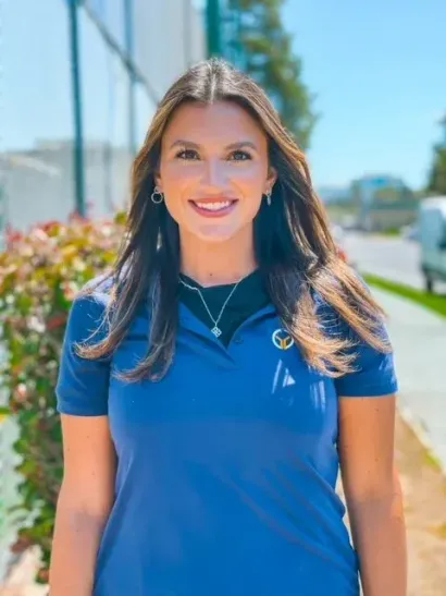 Smiling woman in a blue polo shirt standing outdoors in bright sunlight.