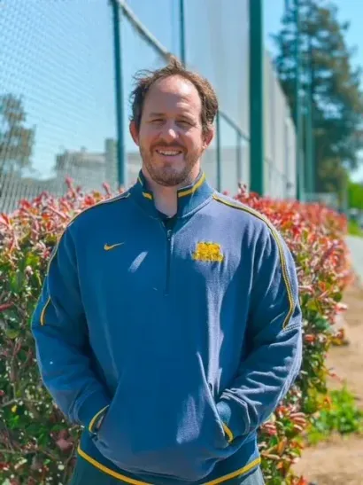 Man standing outdoors in a blue jacket, smiling in front of bushes and a glass building.