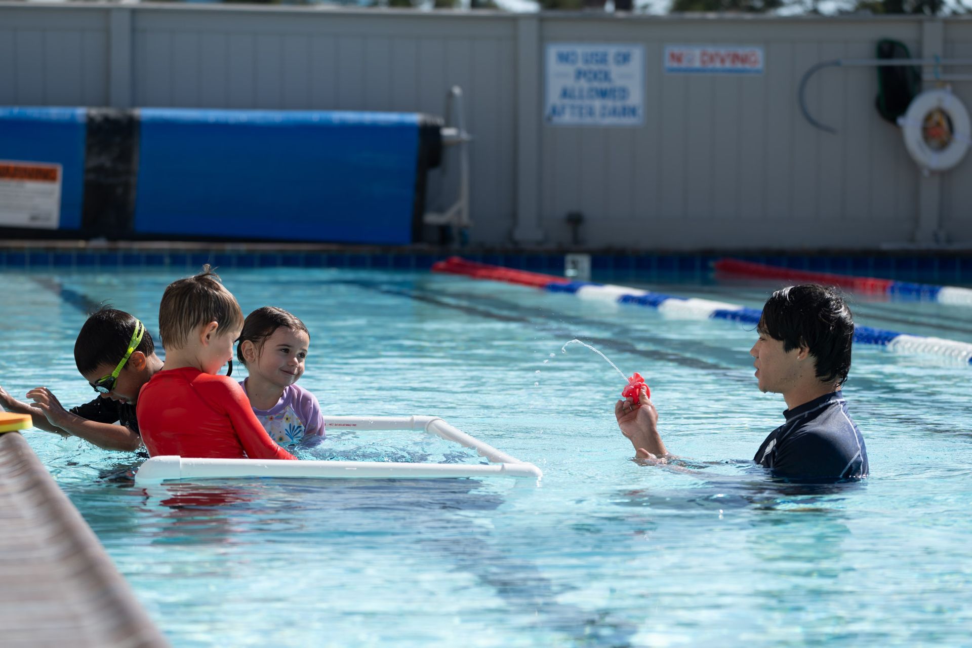 Swimmers in a pool with an instructor holding a red toy, near a floating white frame