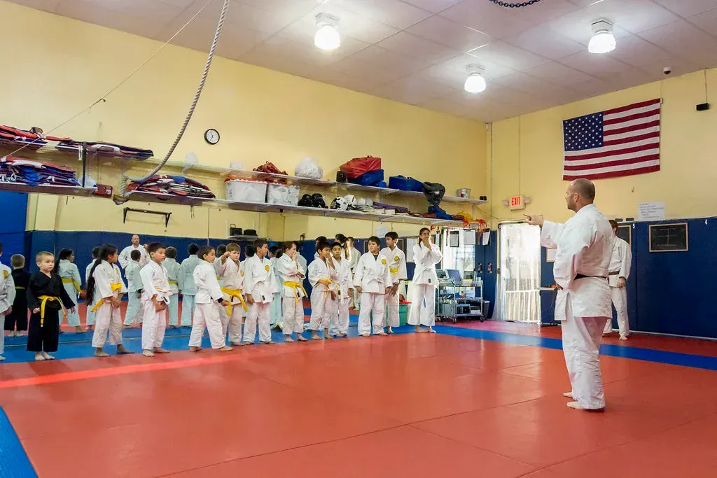 Martial arts class lining up in a dojo, instructor facing students on red mats with an American flag in back.