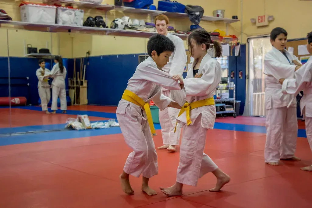 Two children in white judo gis practice grappling on a red mat in a dojo