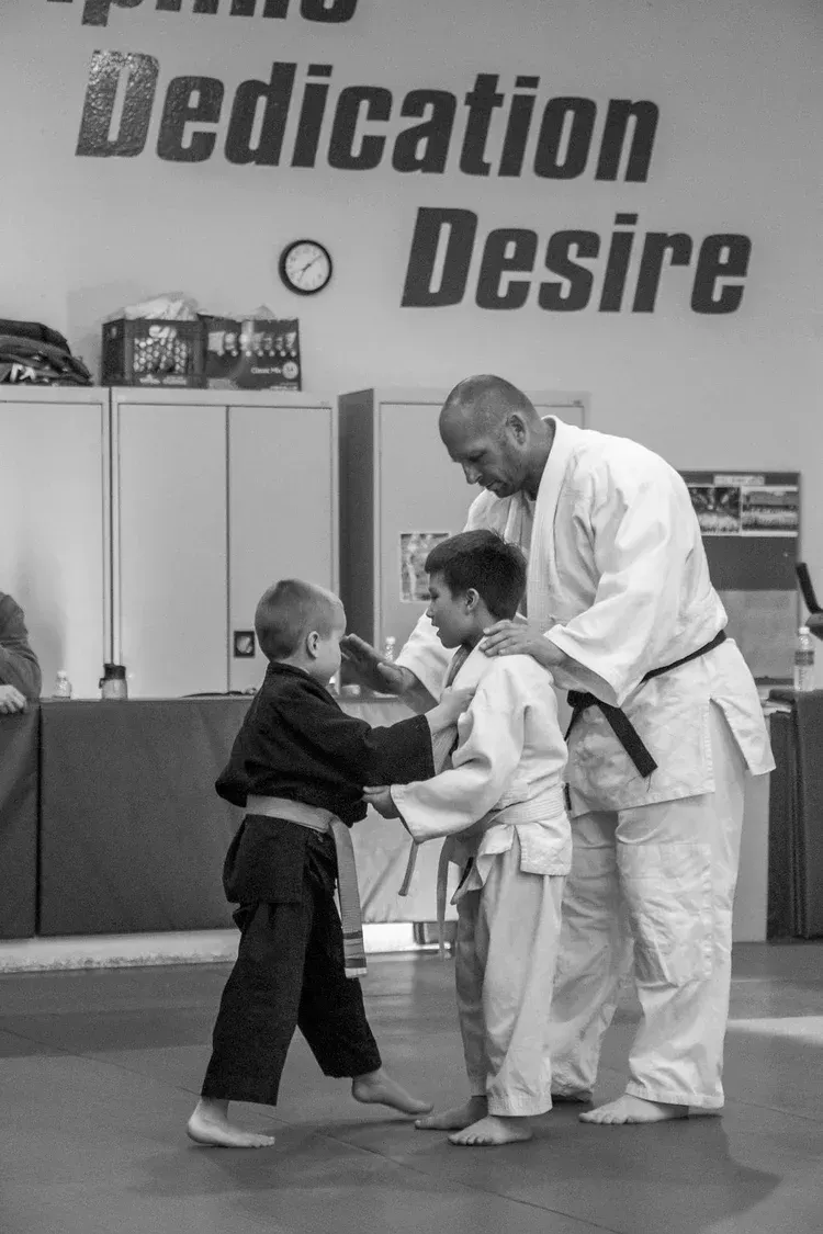 Two martial arts students practicing with an instructor on a mat, with “Dedication Desire” on the wall behind them.