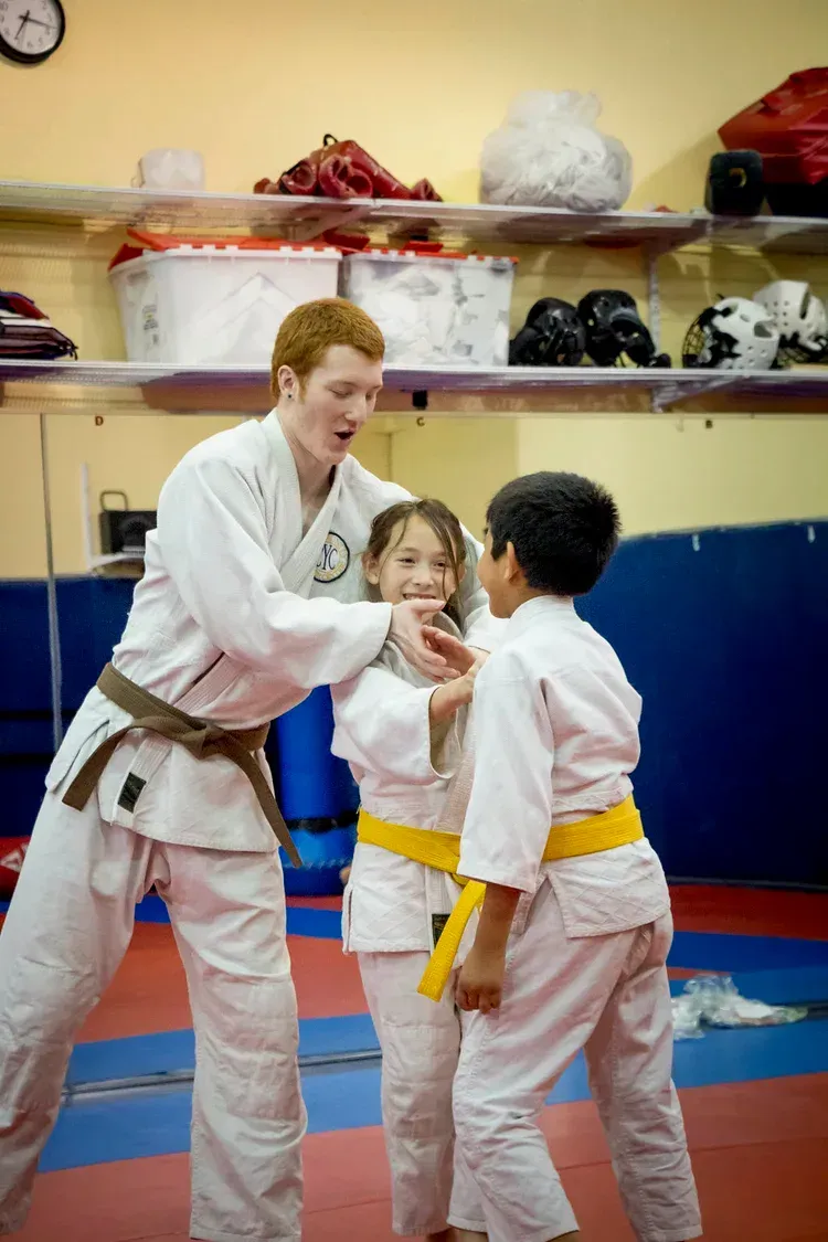 Judo instructor in white gi coaching two students on mats, with shelves of gear in the background