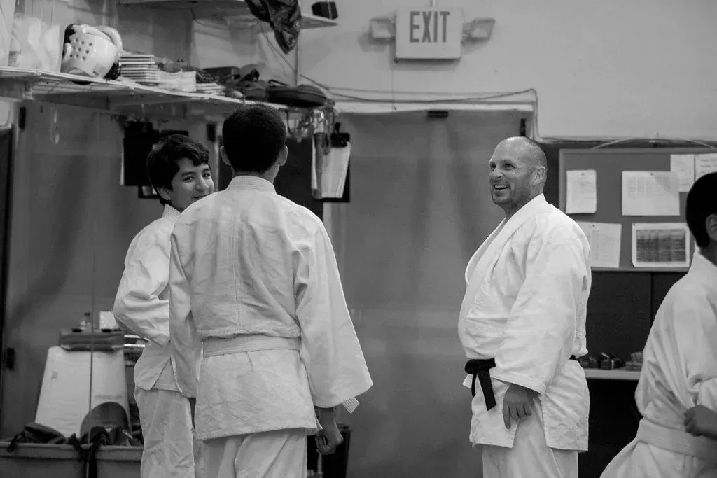Martial arts class in a gym, with students in white uniforms and one instructor smiling near the exit.