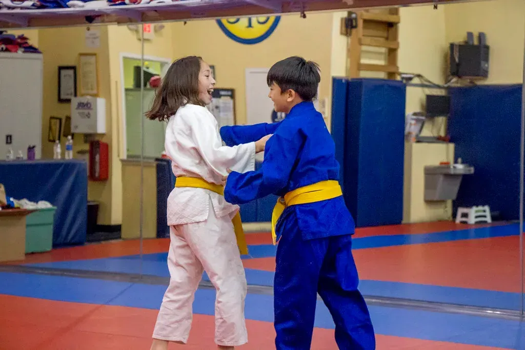 Two children practicing a martial arts hold in a dojo, one in blue and one in white uniforms