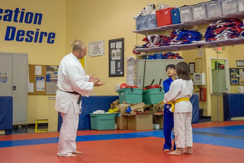 MARTIAL ARTS CLASS: instructor in white gi teaches two students in blue and white gis in a dojo.