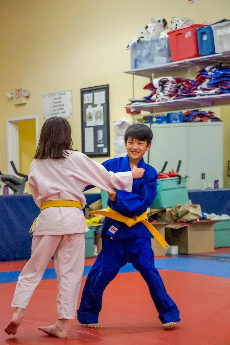 Two kids in martial arts uniforms practicing a partner drill in a dojo with mats and shelves.