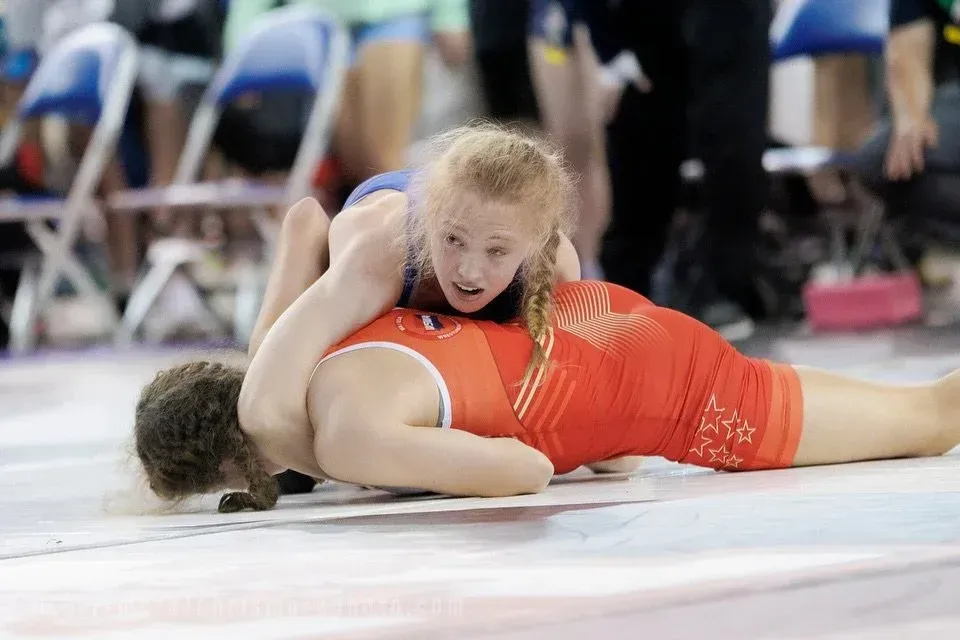 Two wrestlers on a mat, one pinning the other during a match, with spectators in the background
