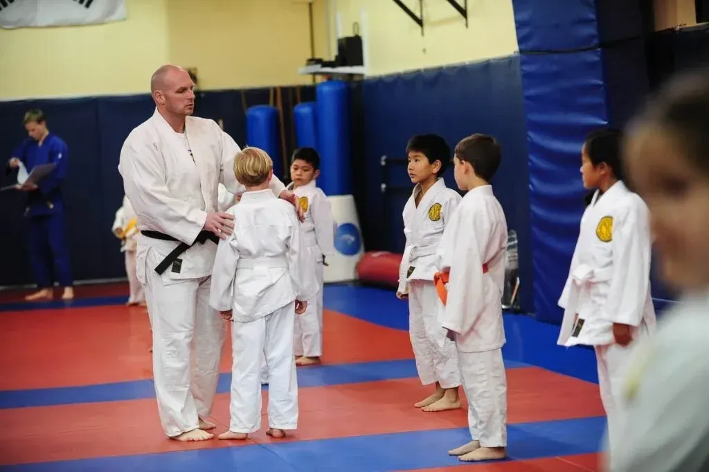 Karate instructor speaking to children in white uniforms on a red-and-blue mat in a gym