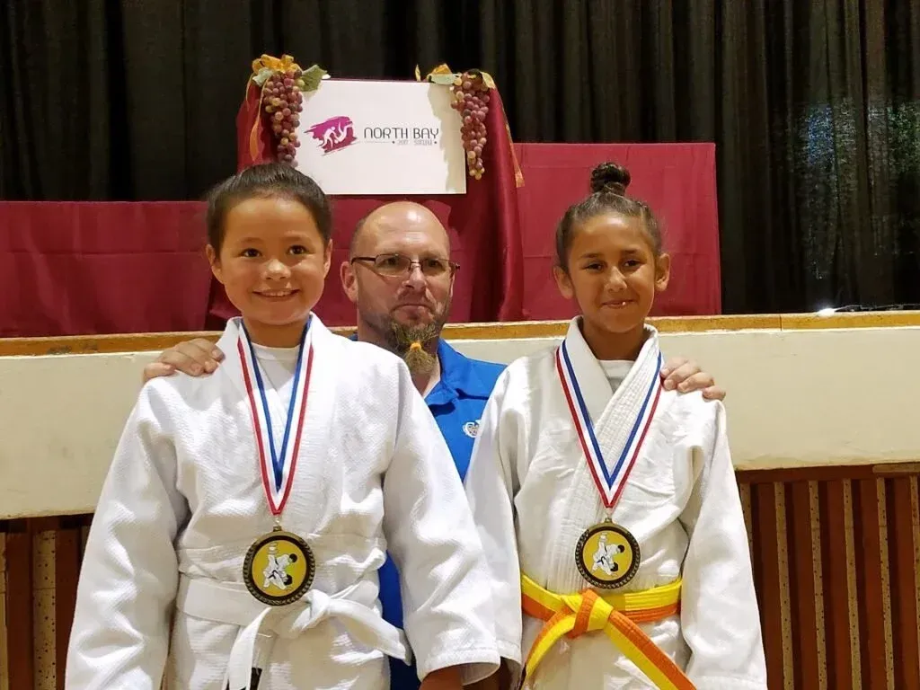 Two children in martial arts uniforms with medals, standing with a man on a stage