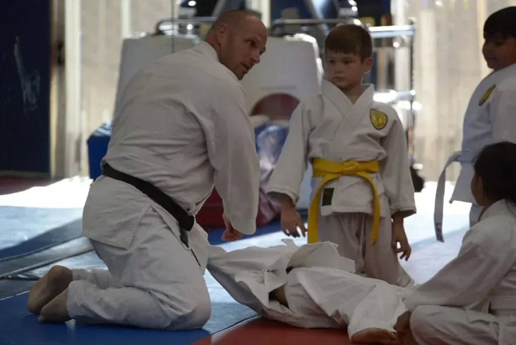Martial arts instructor kneels beside a child in a white gi during a training session on blue mats.