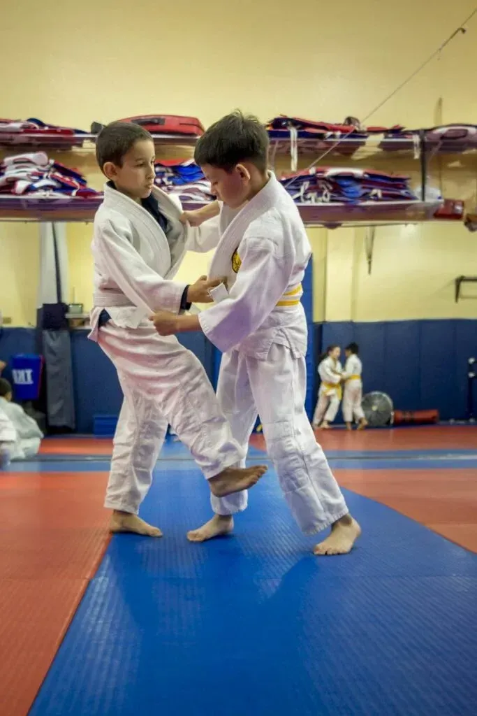 Two martial arts students in white gis practicing grappling on blue mats in a dojo