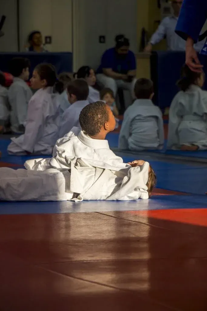 Children in white martial arts uniforms sitting on mats in a gym during a class or event