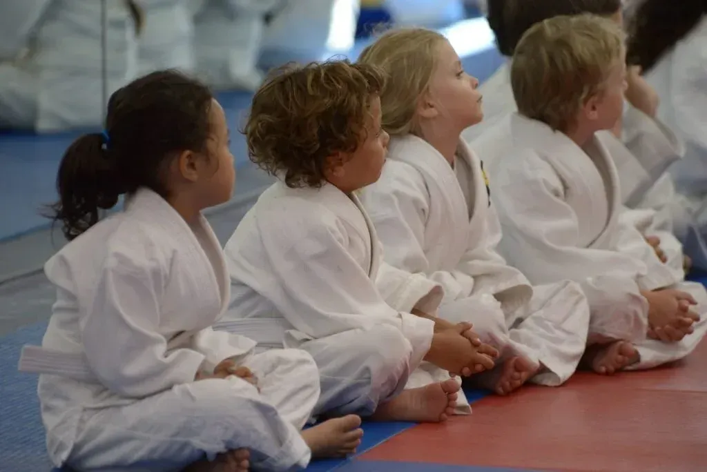 Children in white martial arts uniforms sitting on mats in a dojo, watching attentively
