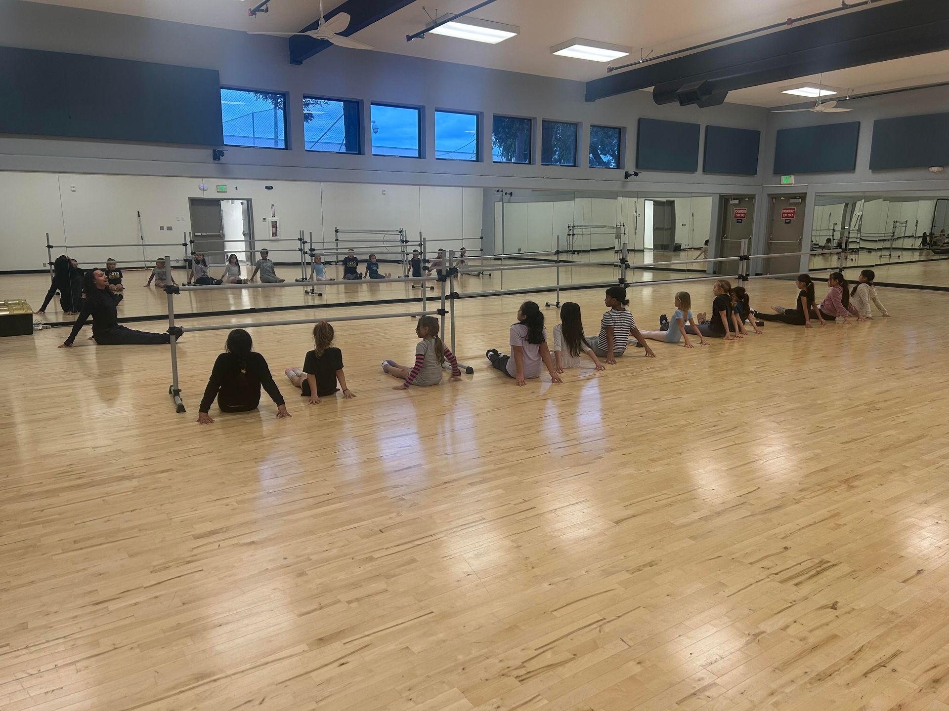 Children seated on a gym floor, facing a mirrored wall in a dance studio