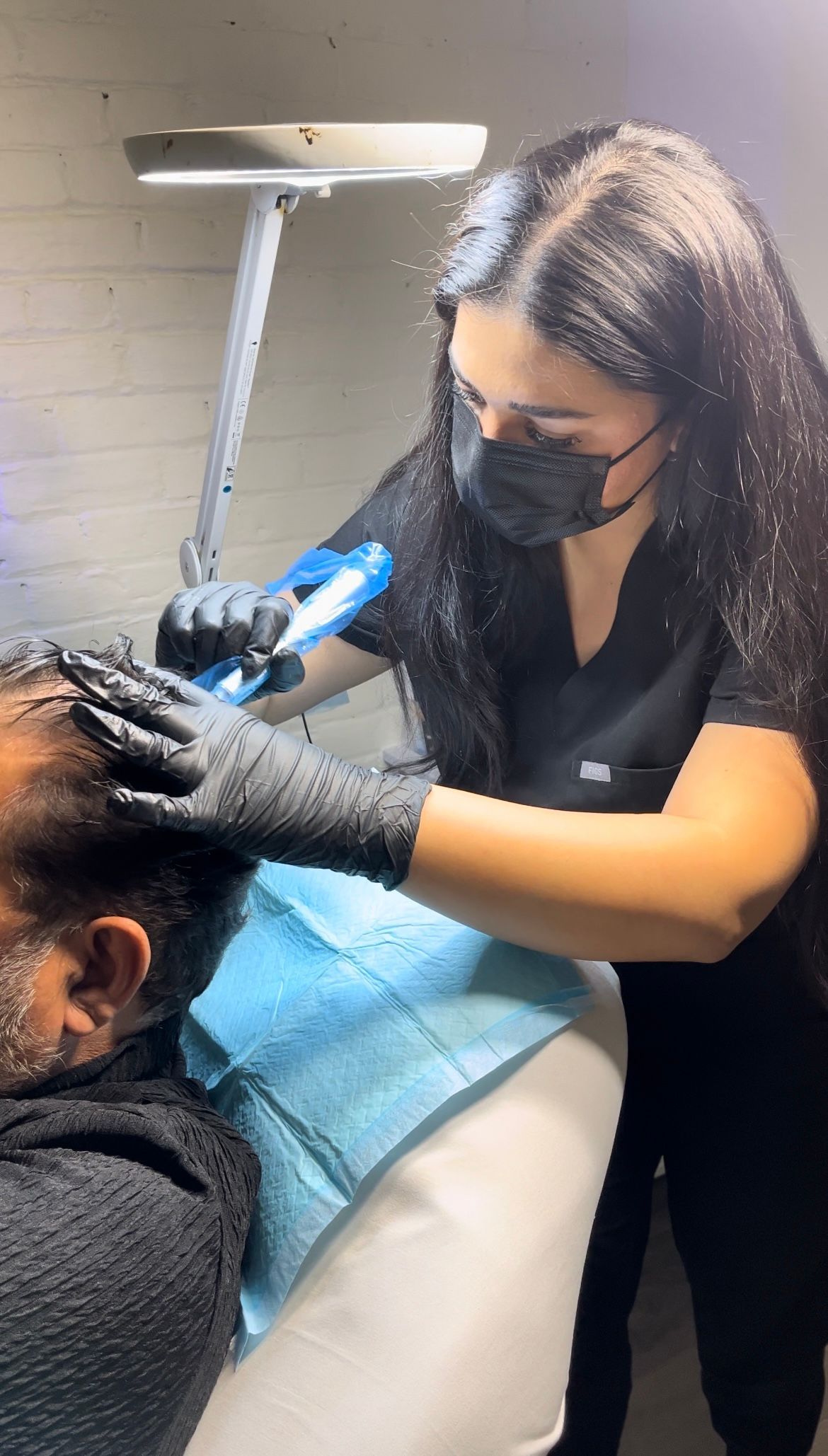 Stylist using blue curling iron on a client’s hair in a salon, wearing black gloves and a mask.