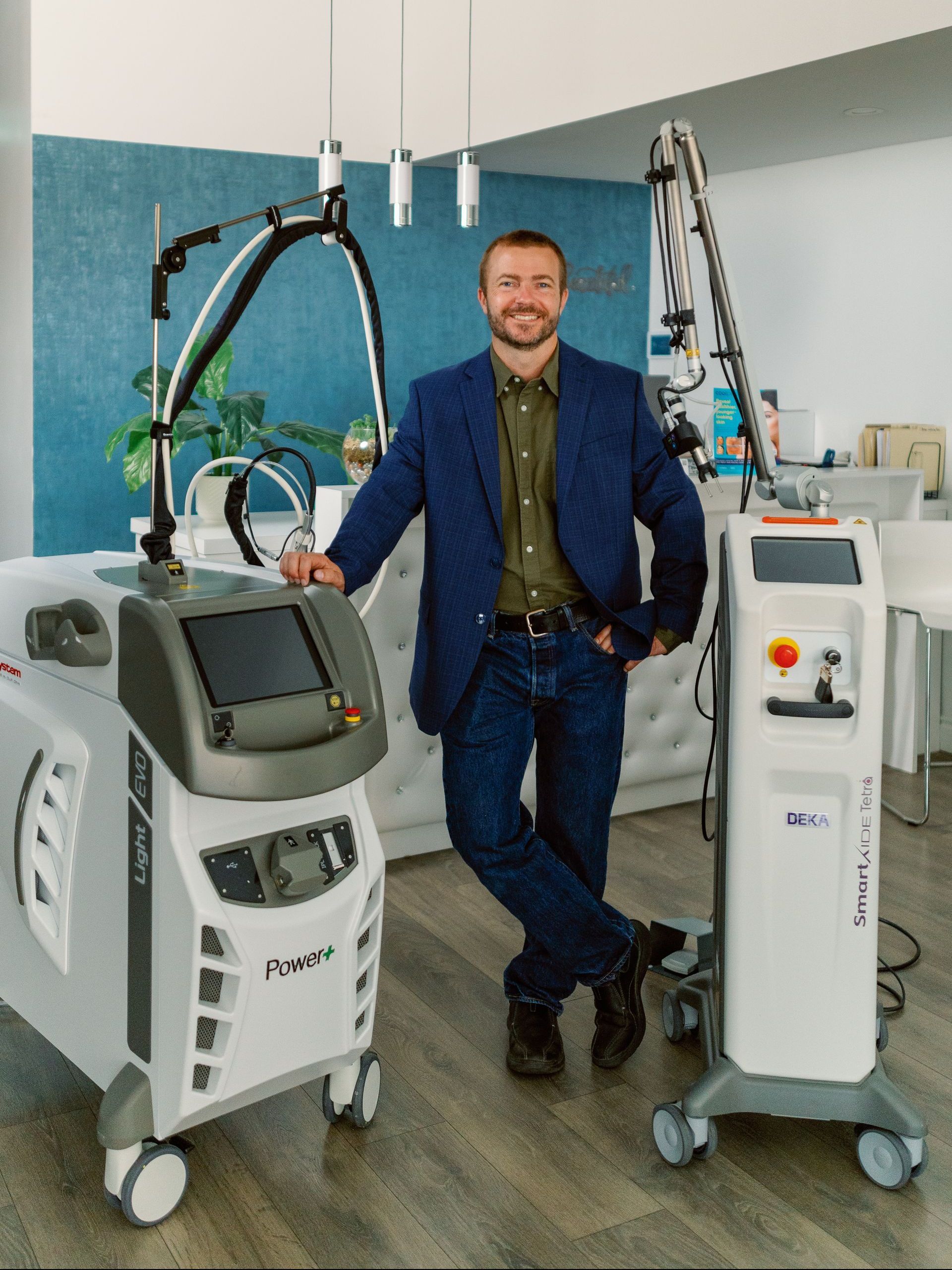 Man standing between two medical machines in a clinical room, smiling in a blue suit.