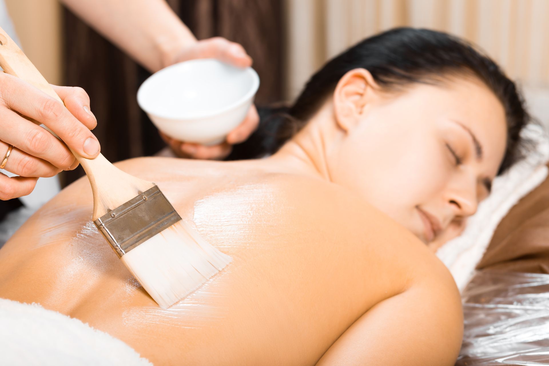 Spa treatment applying lotion to a woman’s back with a brush