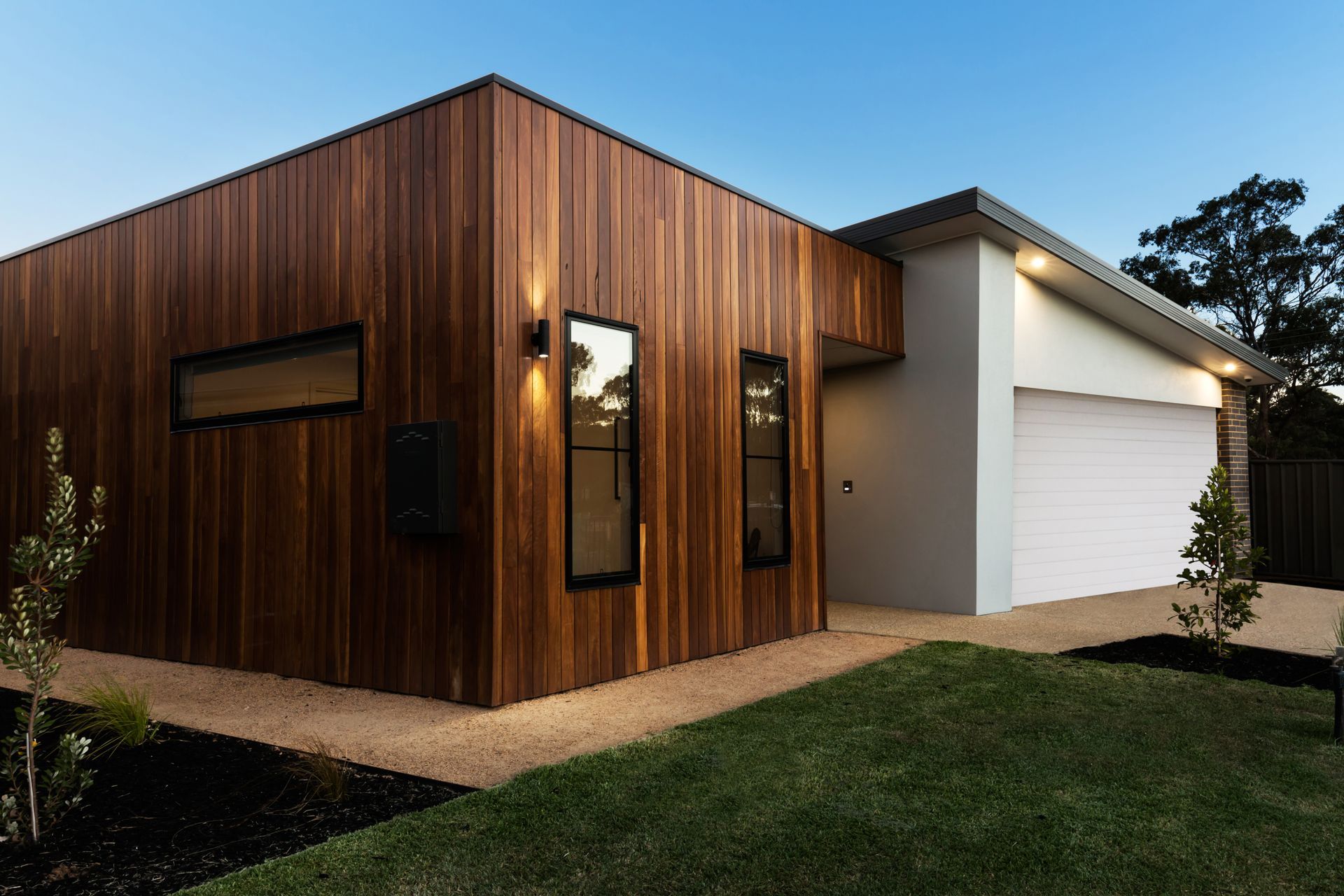 Modern house with wood siding, neutral paint, and a garage door. Small yard with greenery.