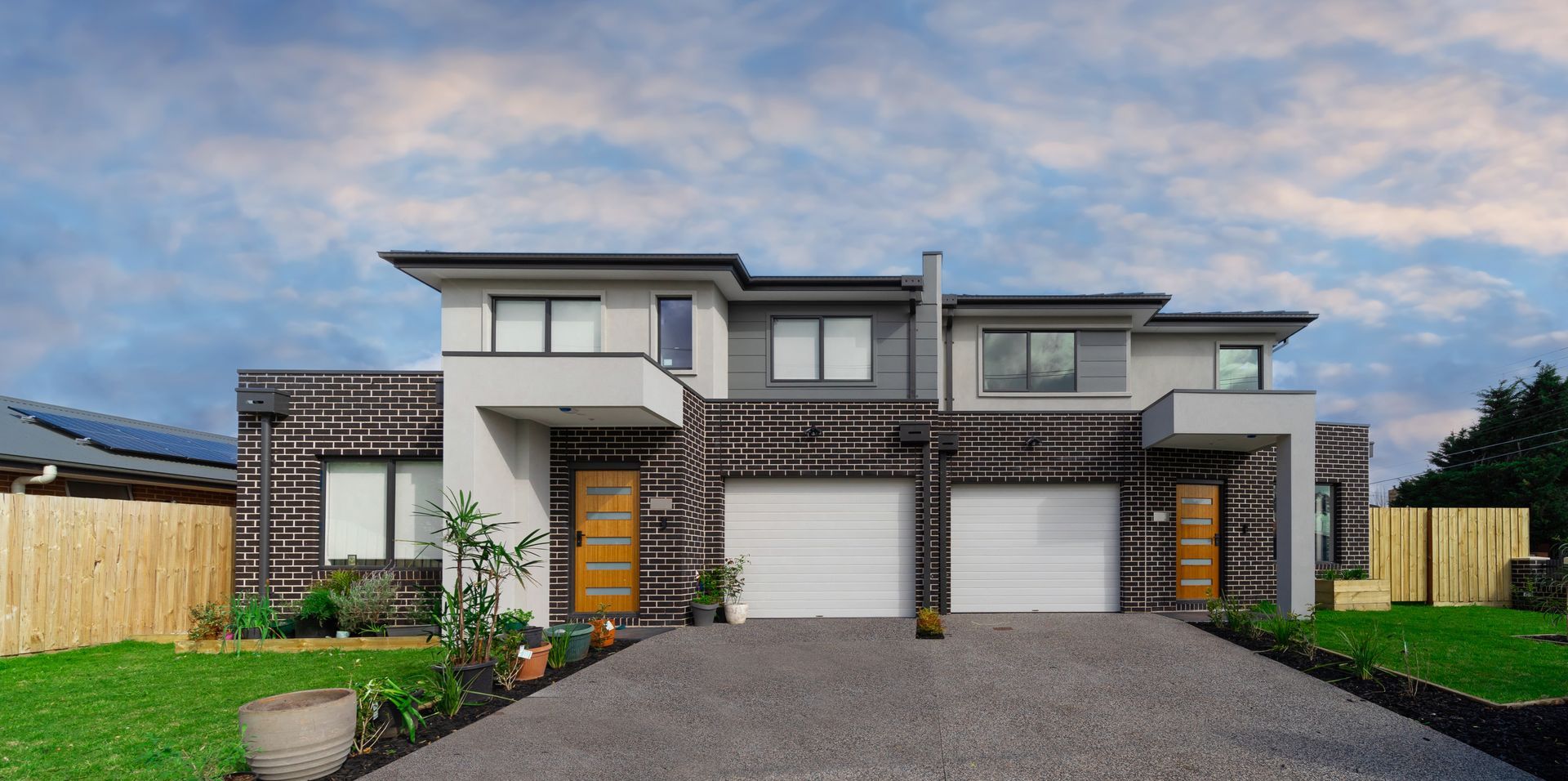 Modern, two-story houses with light gray and dark brown exteriors. Gray driveway, white garage doors, and green lawns.
