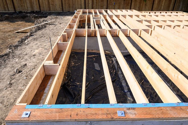 Wooden floor framing under construction, outdoors. Lumber on a concrete base, plastic sheeting visible.