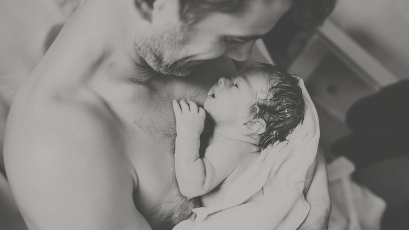 Father gazes lovingly at newborn baby wrapped in a towel against his bare chest. Black and white.