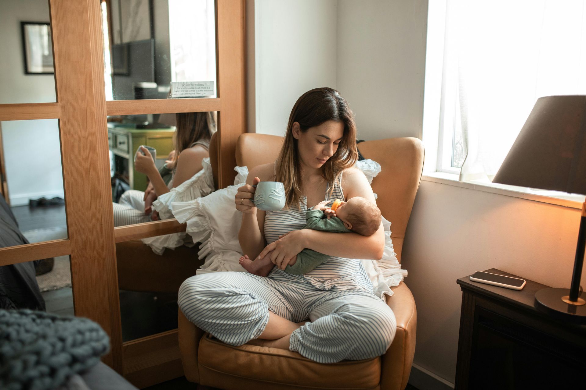 Woman in pajamas holds newborn, drinking from a mug, seated in a leather chair near a window.