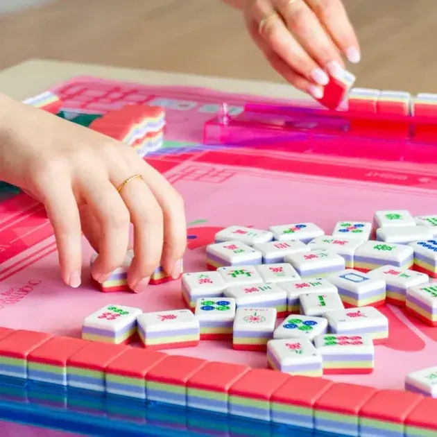 Hands playing a pink mahjong game, arranging tiles on a table.