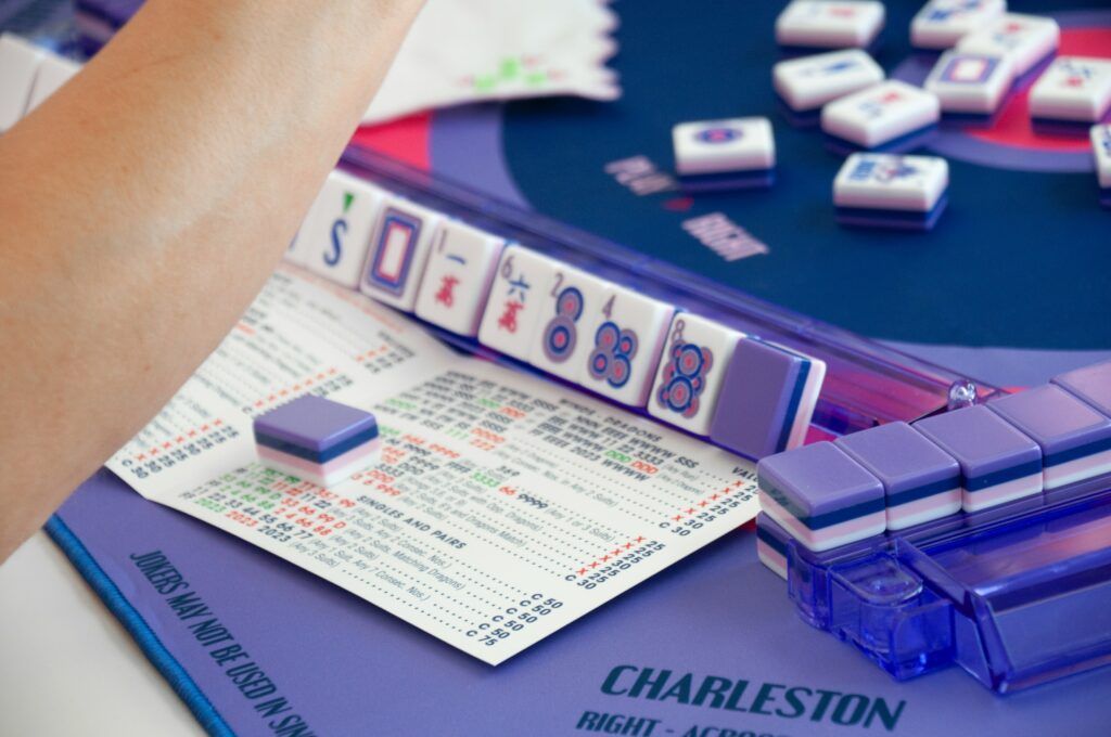 Person playing mahjong; tiles, board, score sheet visible, setting indoors, mostly purple and white.