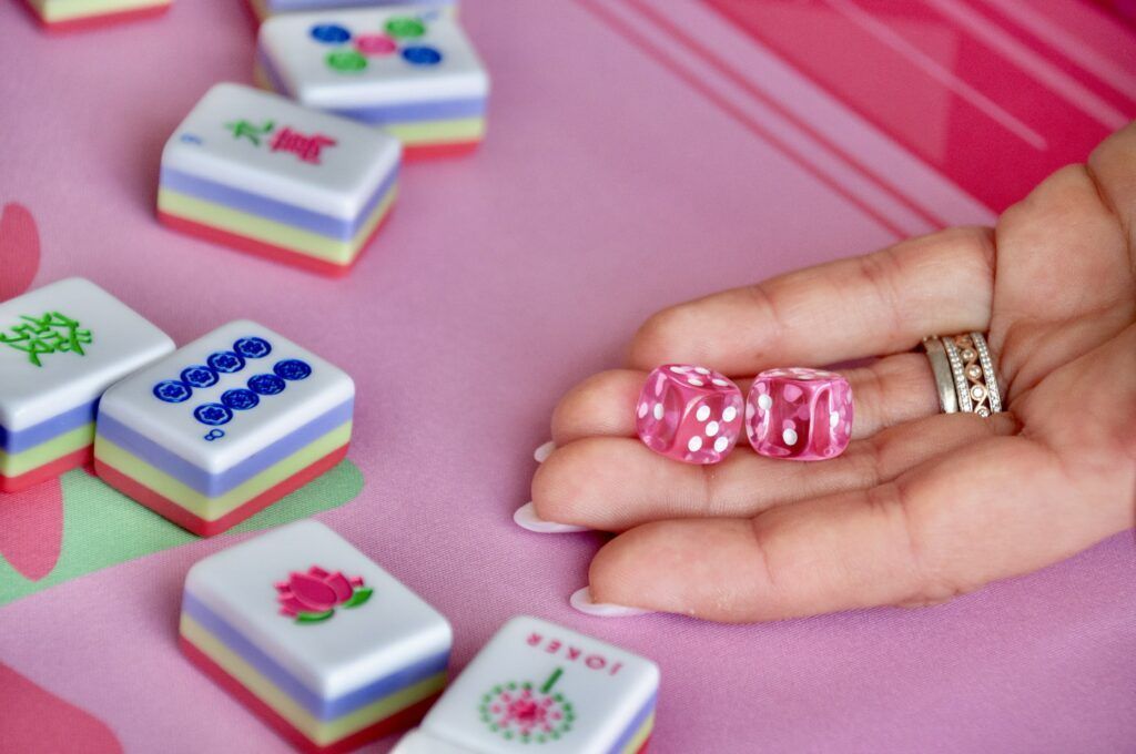 Hand holding pink dice next to Mahjong tiles on a pink surface.